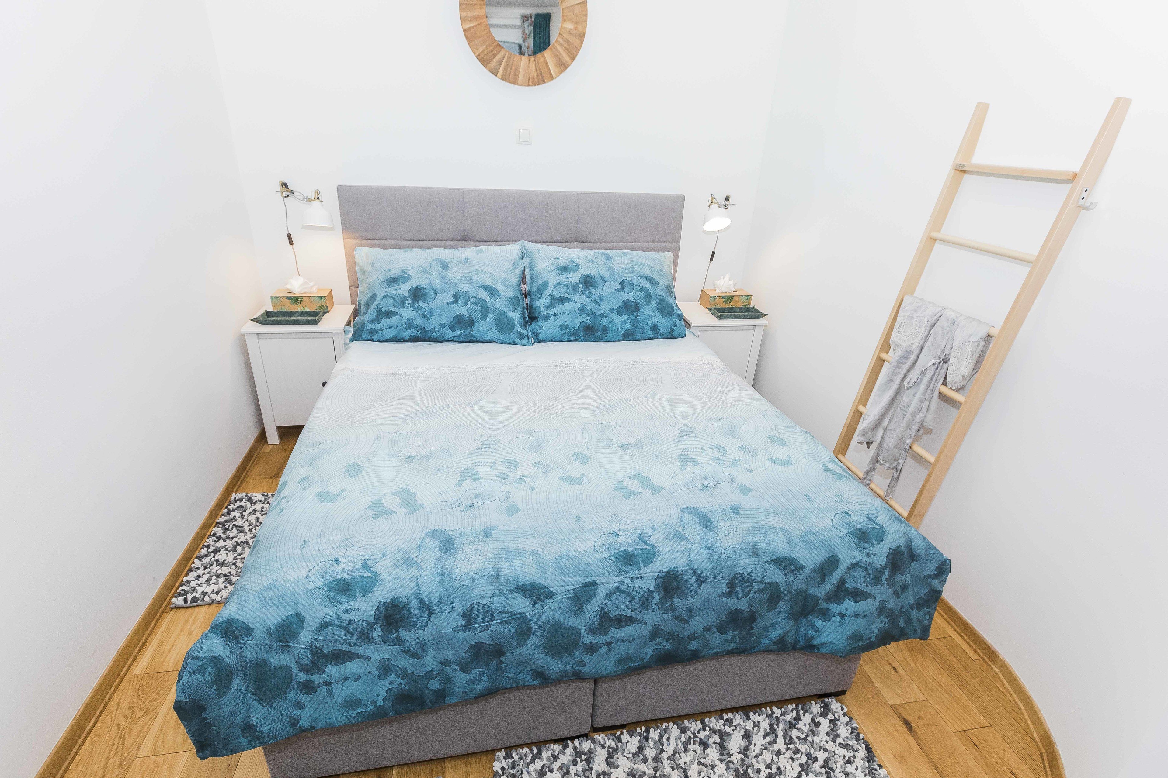 A bed with a blue patterned bedspread, two bedside lamps, a wooden ladder, and a small round mirror on the wall.