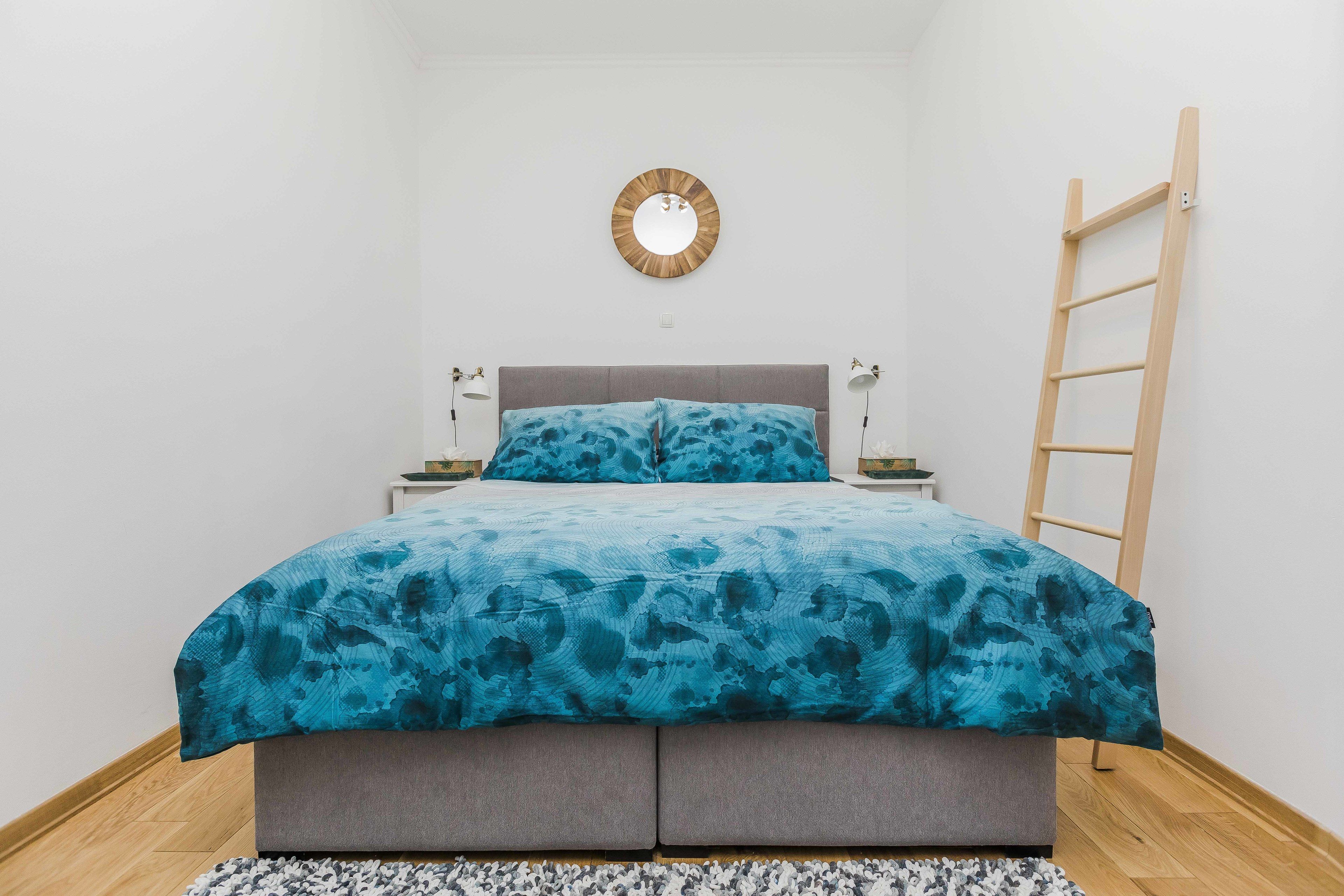 Quarto com cama, espelho redondo, escada de madeira e luminárias de cabeceira.