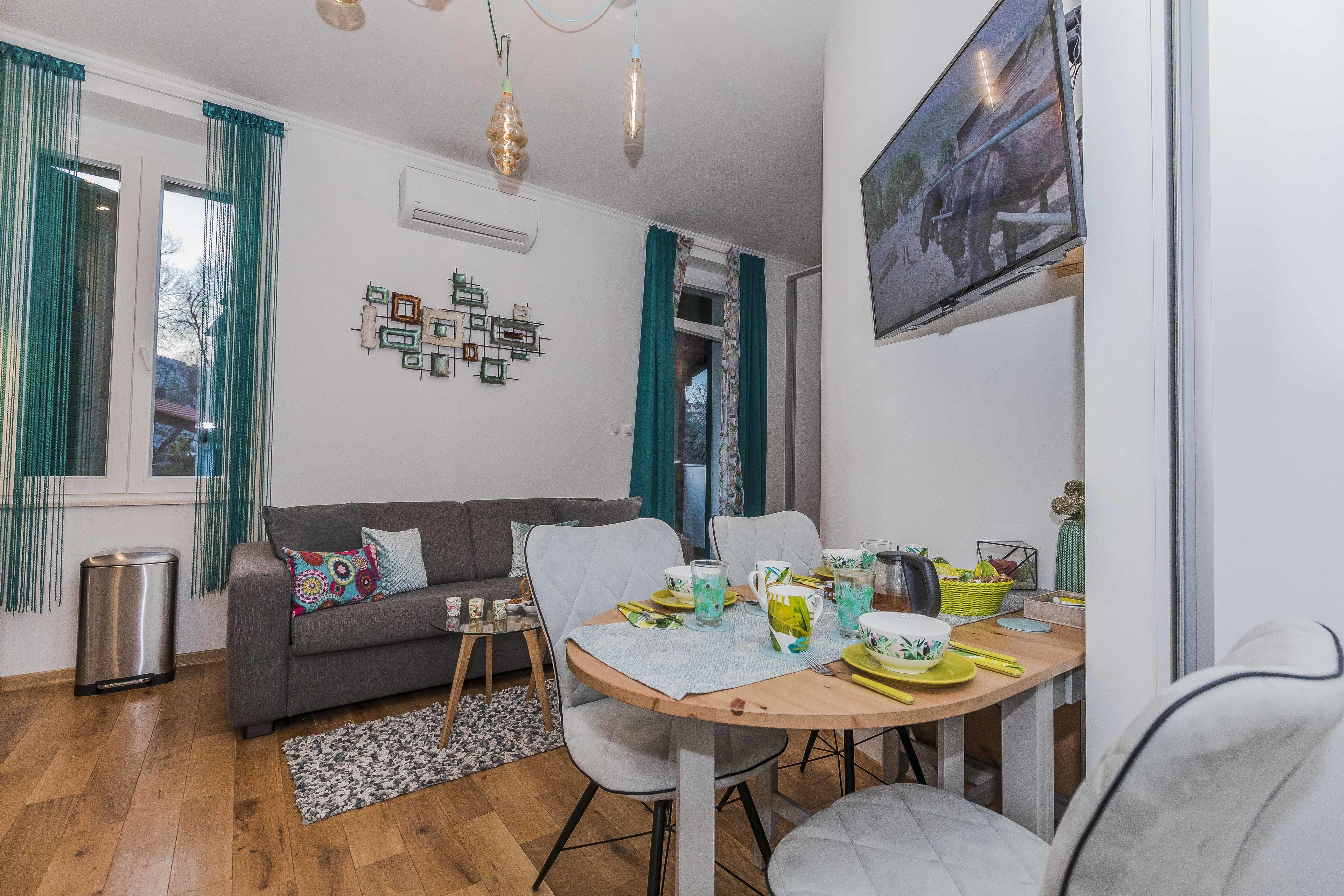 A modern living room with a grey sofa, a dining table set with yellow plates and glasses, and a TV mounted on the wall.