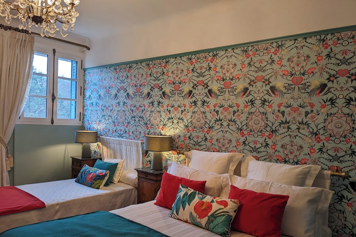 Ein Schlafzimmer mit zwei Betten, Blumenmuster-Tapete, einem Chandelier und einem Fenster mit Vorhängen.