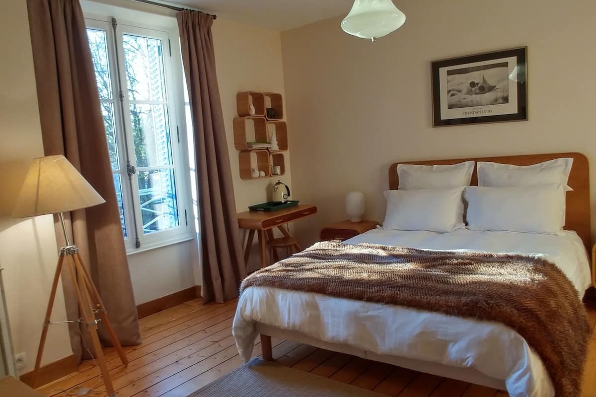 Ein Schlafzimmer mit einem hölzernen Bett, einem Nachttisch, einer Lampe, einem Fenster mit Vorhängen, einem Regal mit Dekorationen und einem gerahmten Bild an der Wand.
