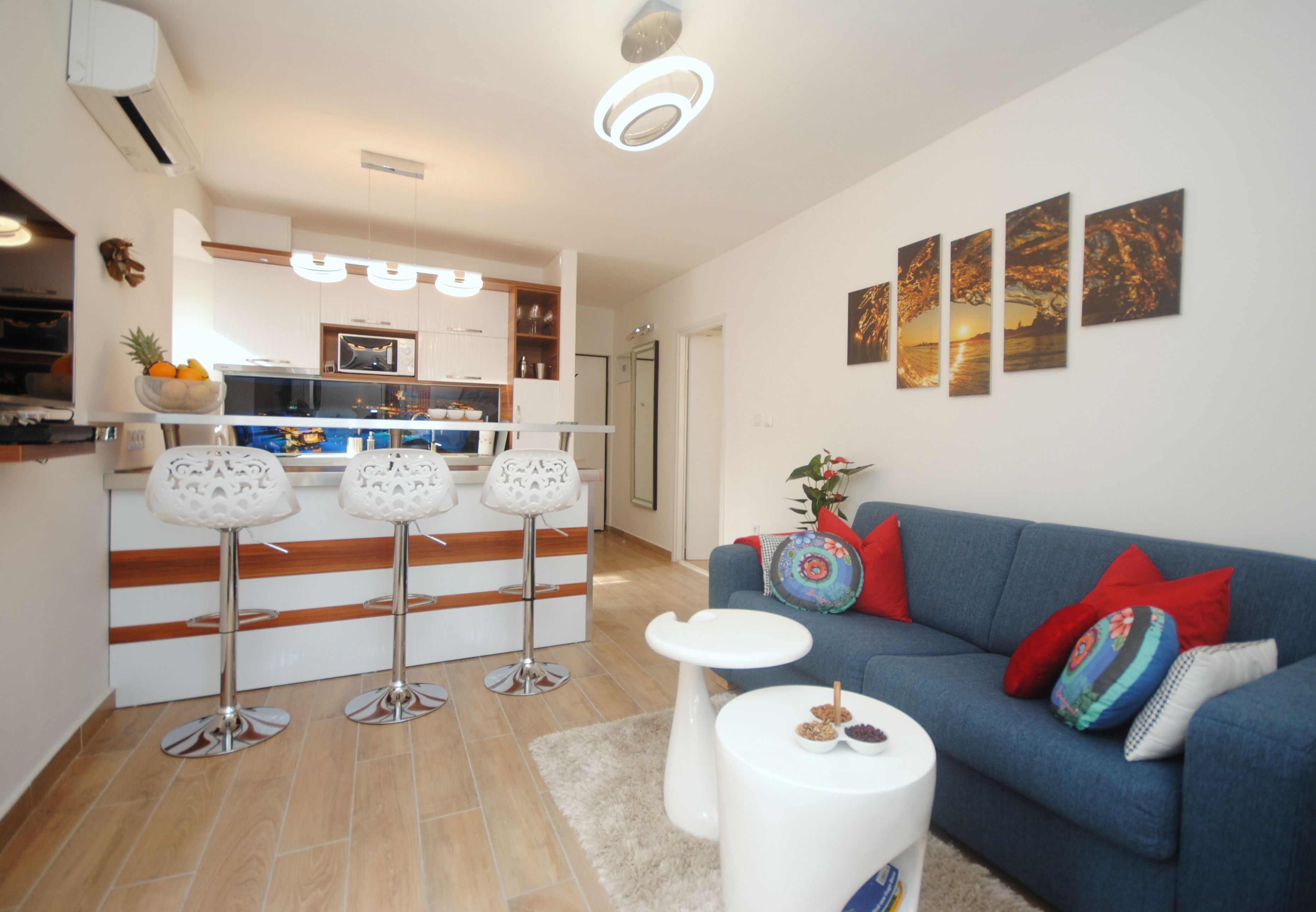 A modern living room with a blue sofa, white coffee table, and a kitchen area with a fruit bowl and bar stools.