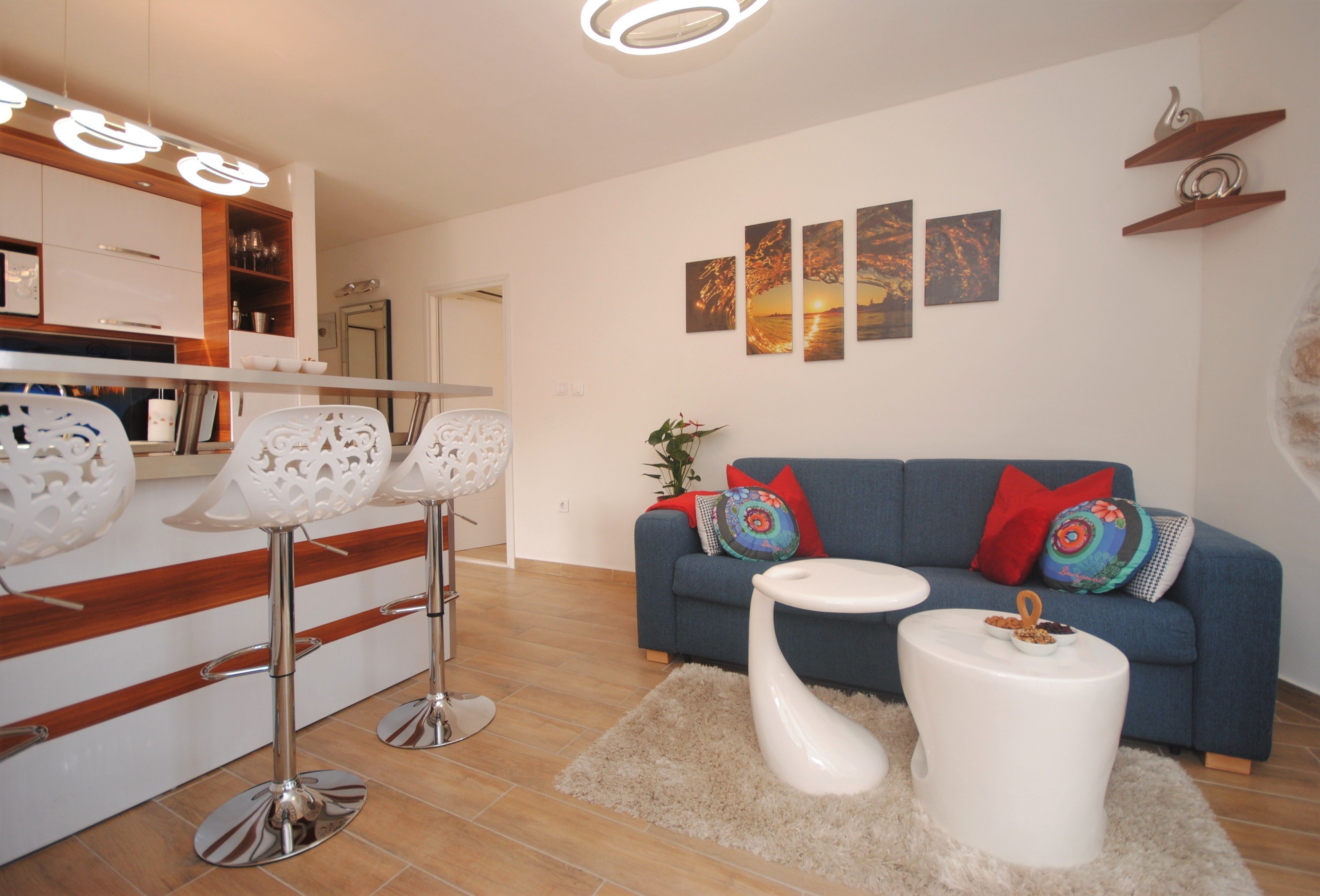Un salón moderno con un sofá azul, una mesa de centro blanca y taburetes de bar. Hay estanterías montadas en la pared y un cuadro.