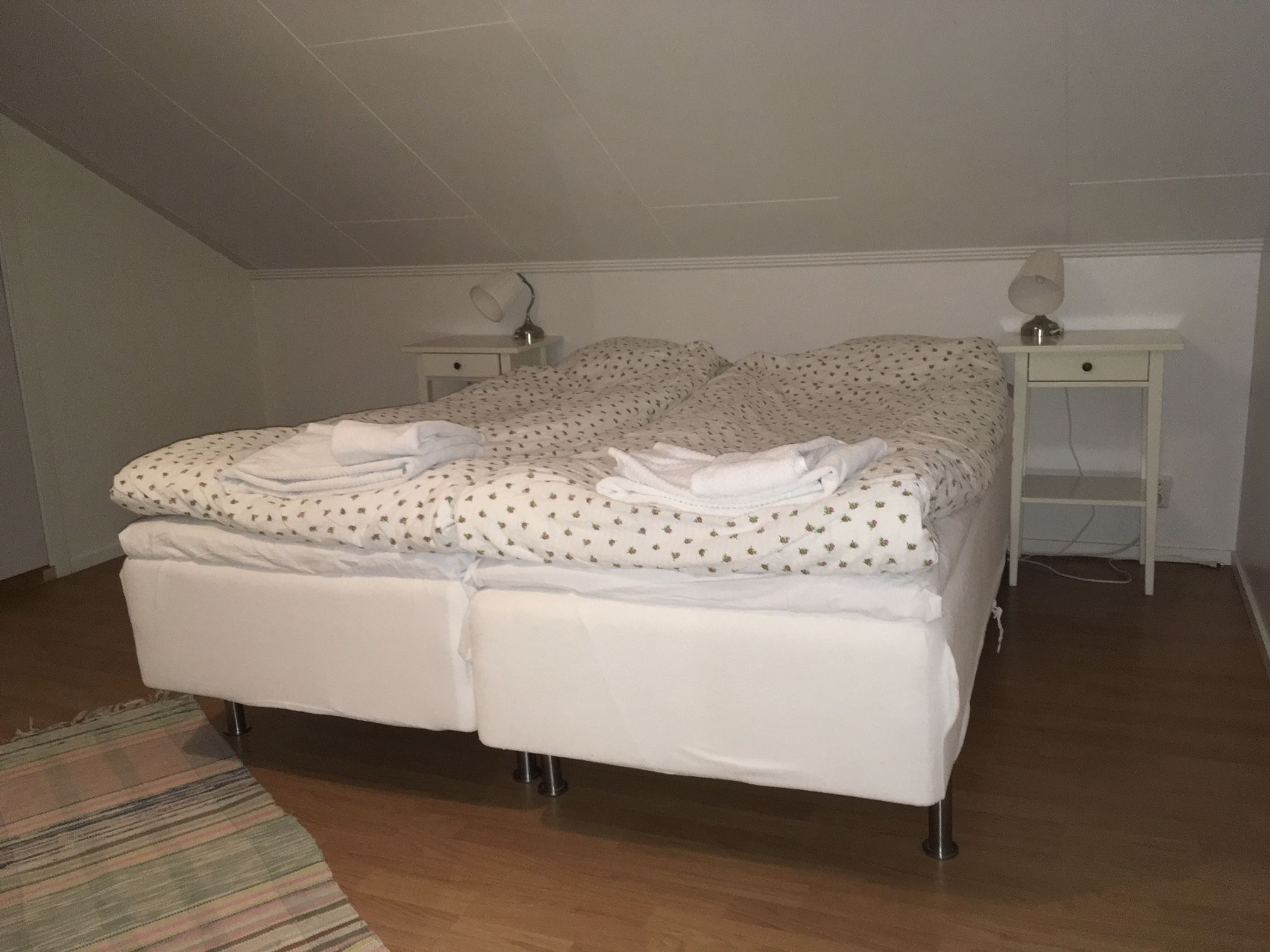 Una cama bien hecha con ropa de cama blanca y varias toallas dobladas, una mesita de noche con una lámpara, y un suelo de madera.