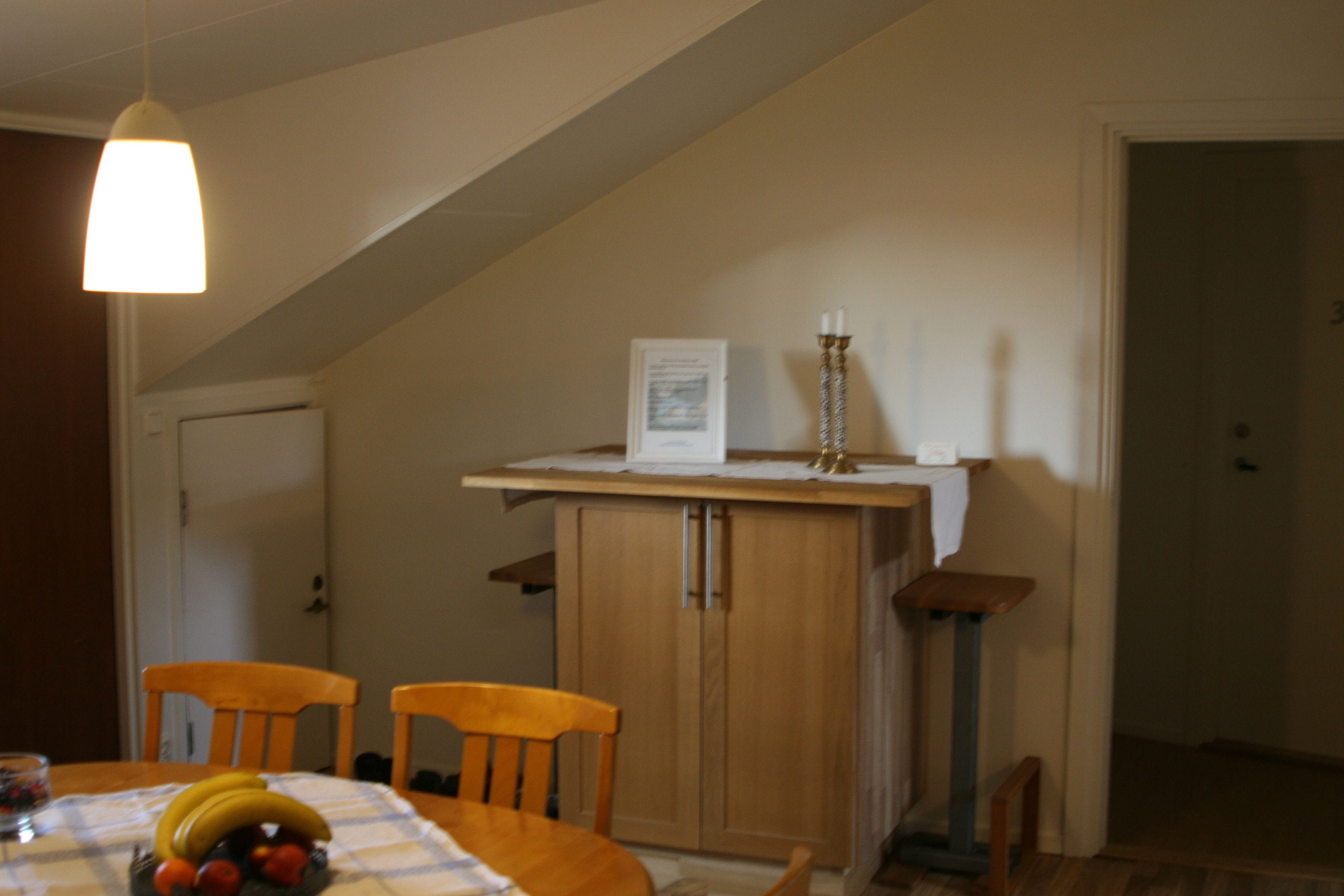 A dining area with a wooden table, two chairs, a hanging light fixture, a cabinet, and a door in the background.