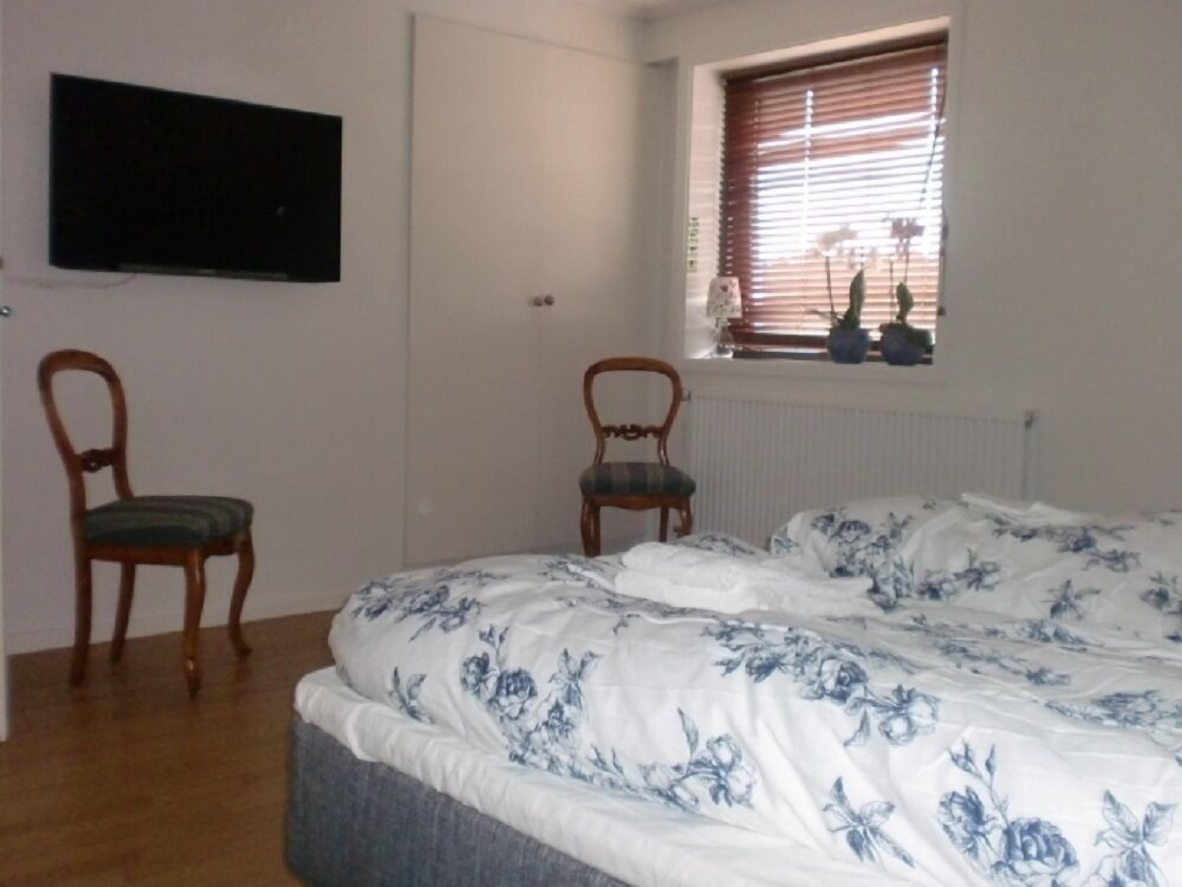 Un dormitorio con una cama, dos sillas de madera, un televisor en la pared y una ventana con persianas.