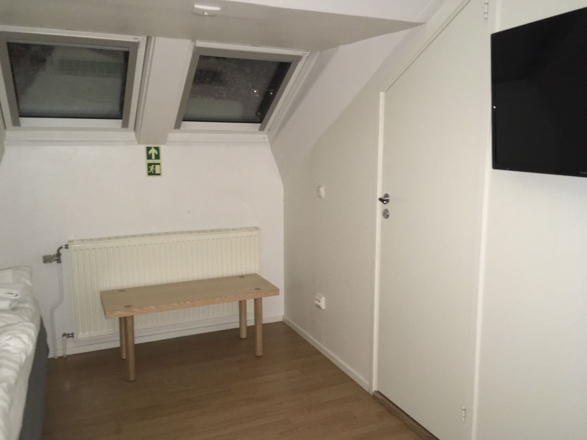 Ein Zimmer mit schrägen Wänden, zwei Fenstern, einem Heizkörper, einer Holzbank und einer Tür.
