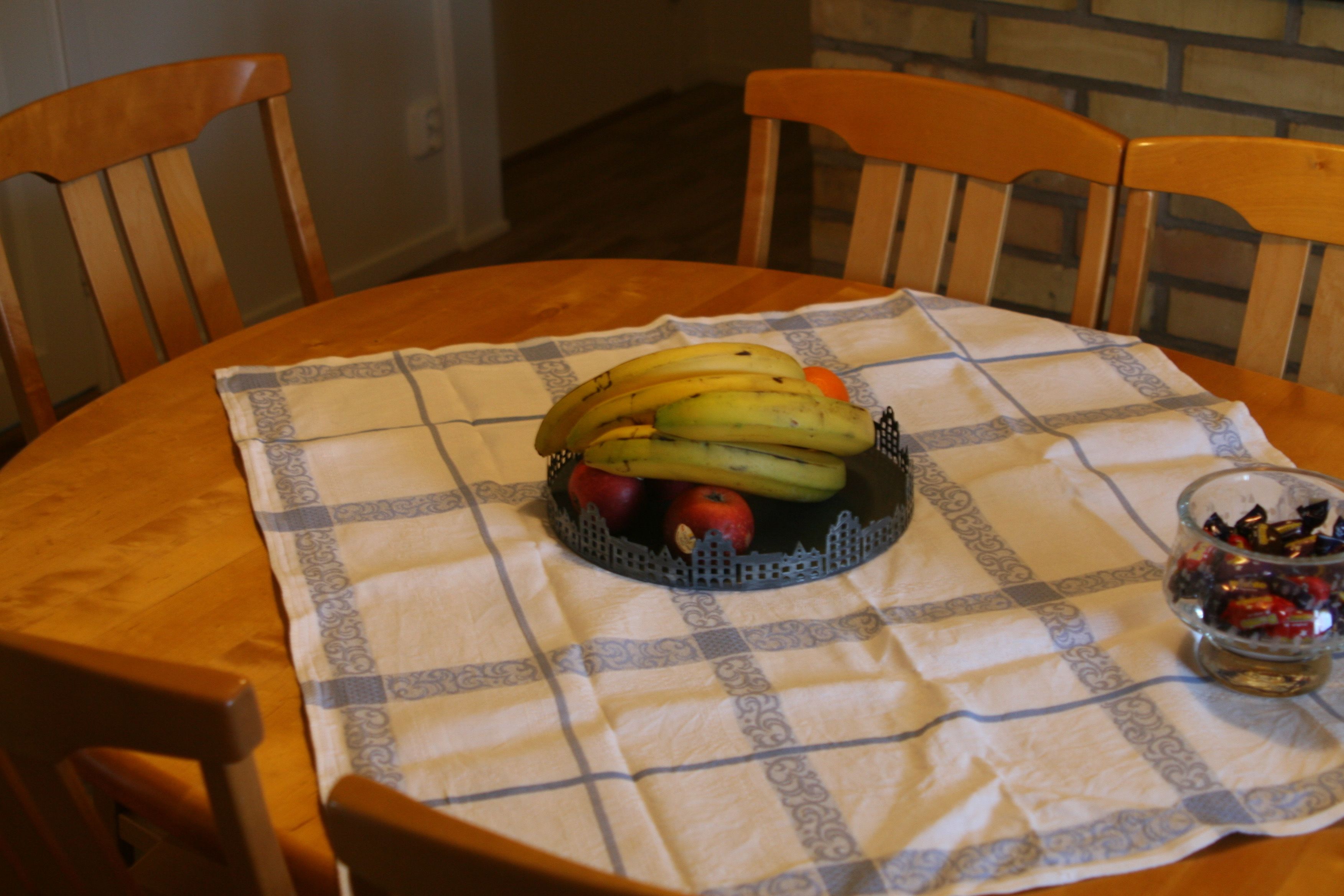 Une table ronde en bois, recouverte d’une nappe à carreaux, est entourée de chaises en bois. Un panier de fruits contenant des bananes et des pommes est posé sur la table.