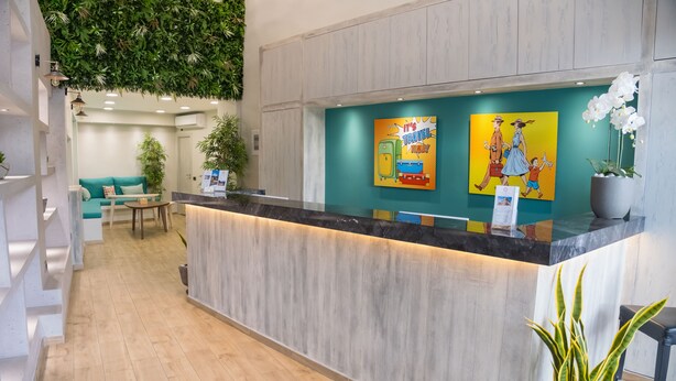 A reception area with a wooden reception desk, a teal wall with travel-themed artwork, and a green hanging plant. - Connect Suites (Athens)