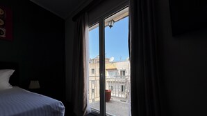 A hotel room with a bed, a lamp, and a view of buildings through a window. - Connect Suites (Athens)