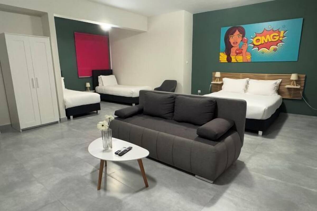 Habitació d'hotel amb un sofà gris, dos llits individuals, una taula blanca amb un ram i un quadre a la paret.