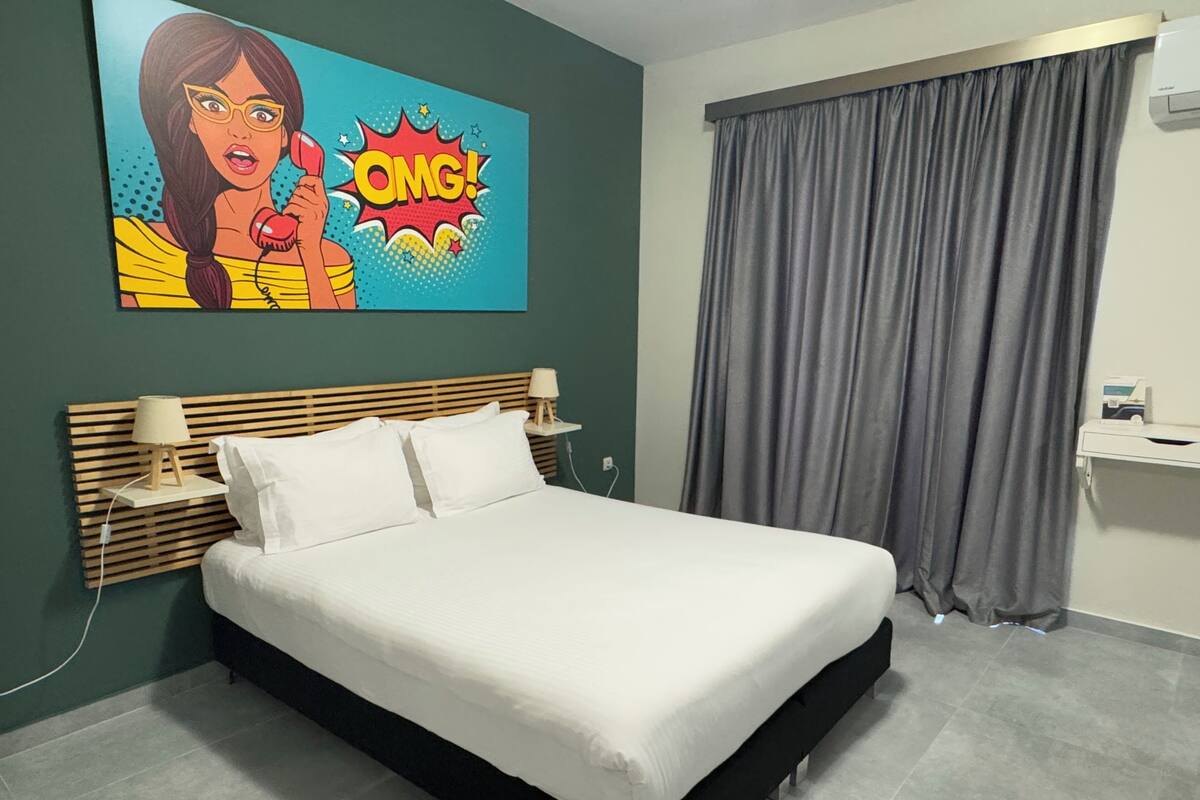Una habitació d'hotel moderna amb un llit gran, dues taules de nit amb làmpades, una cortina gris i una obra d'art colorida a la paret.
