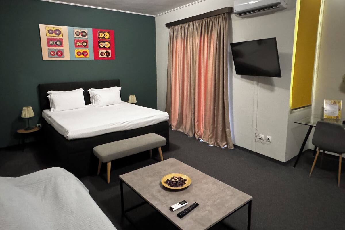 Habitació d'hotel amb un llit, una panxa, una taula petita amb un bol i un comandament, una televisió i una paret amb obra d'art.