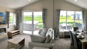 A spacious living room with a sofa, a wooden TV stand, and a dining area with a table set for two. - Newperran Holiday Resort (Truro)