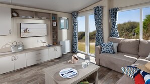 A modern living room with a sofa, coffee table, and a television. There is a window with curtains, a bookshelf, and a view of the outdoors. - Newperran Holiday Resort (Truro)
