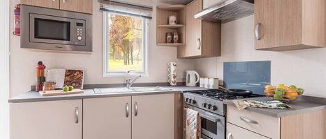 A compact kitchen with a microwave, stove, oven, and sink. There is a window with a view of trees, and a small shelf with jars.