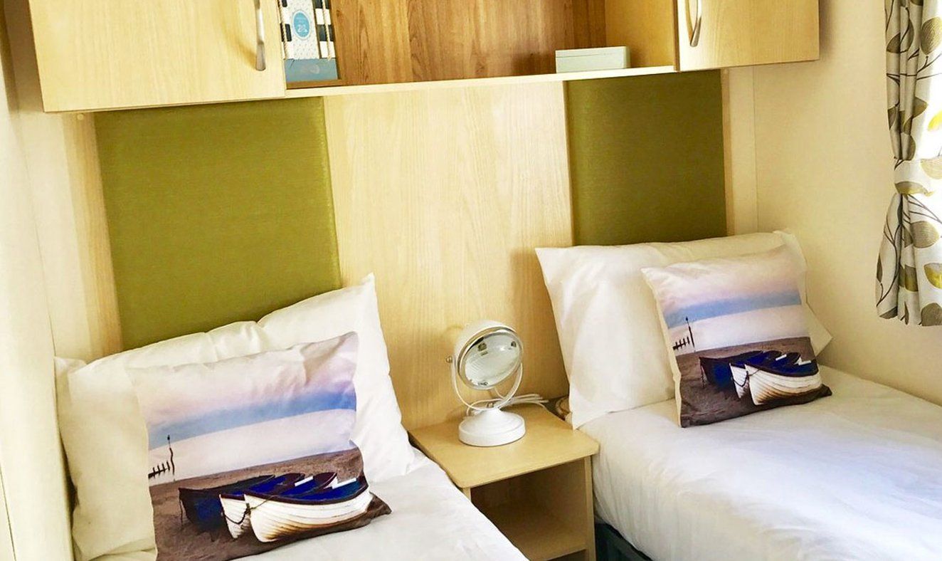 A hotel room with two beds, each with a pillow featuring a boat and sea scene, a bedside table with a mirror, and a small shelf above the beds.