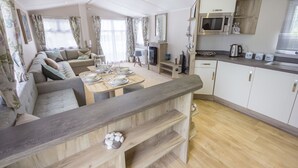 A modern, well-lit living room with a sofa, dining table, and kitchen area. - The Lakes Rookley Holiday Park (Ventnor)