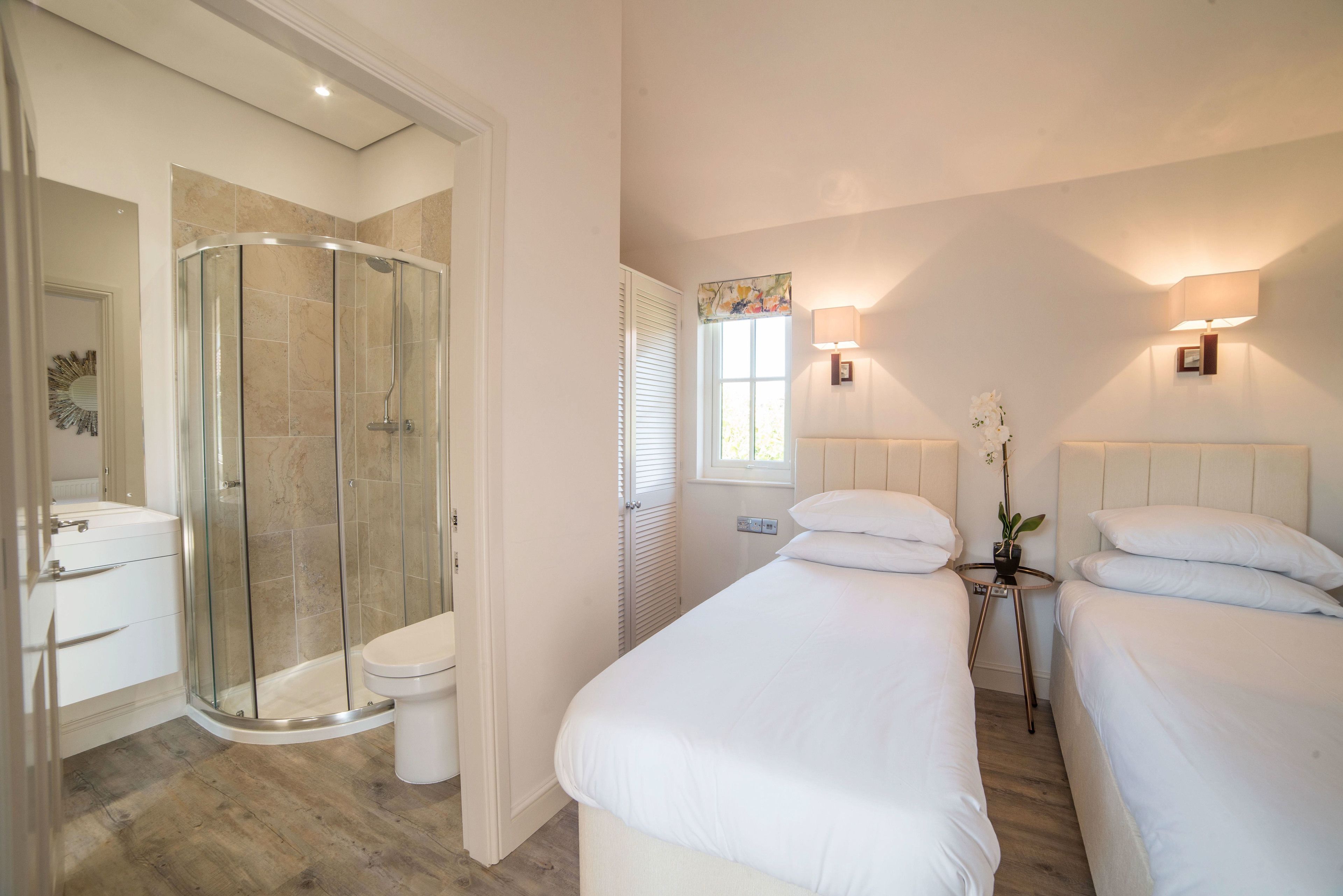 A hotel room with a glass-enclosed shower, a white toilet, a bed with white bedding, and a bedside table with a lamp and a potted plant.