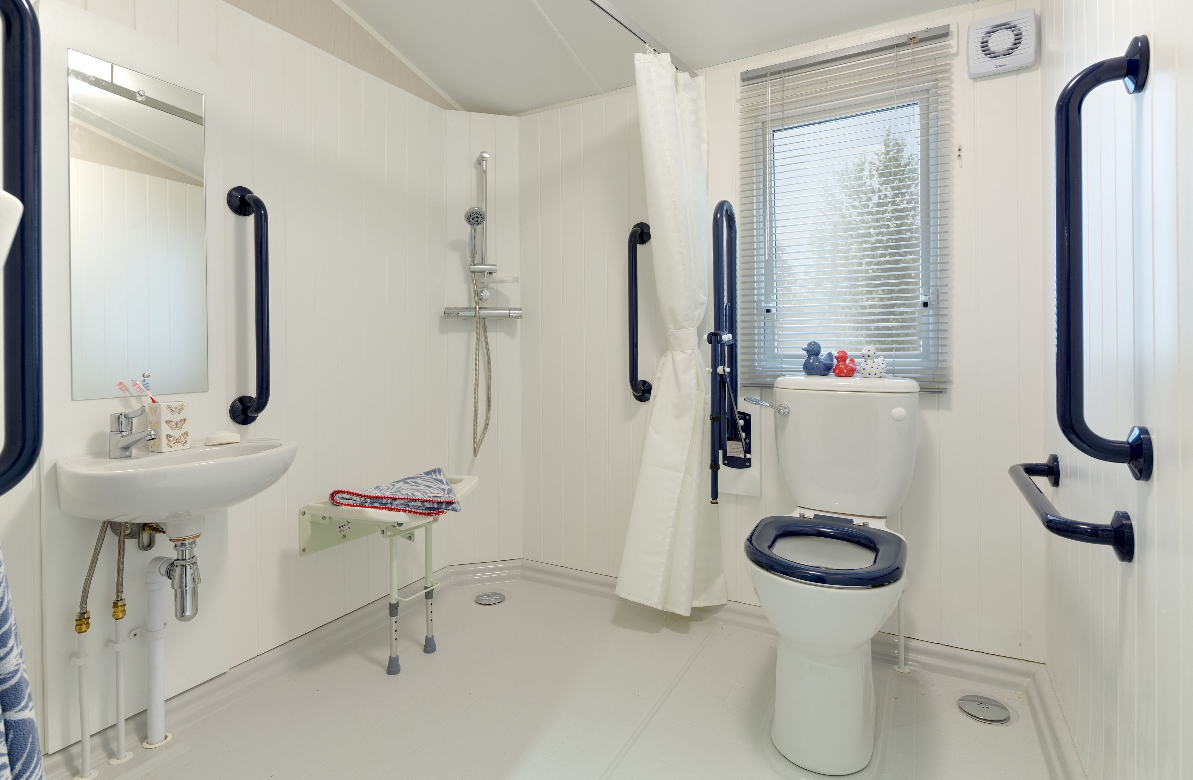 Une salle de bain avec un lavabo, des toilettes et une douche, tous de couleur blanche. Elle est équipée de barres d'appui et d'une fenêtre avec des stores.