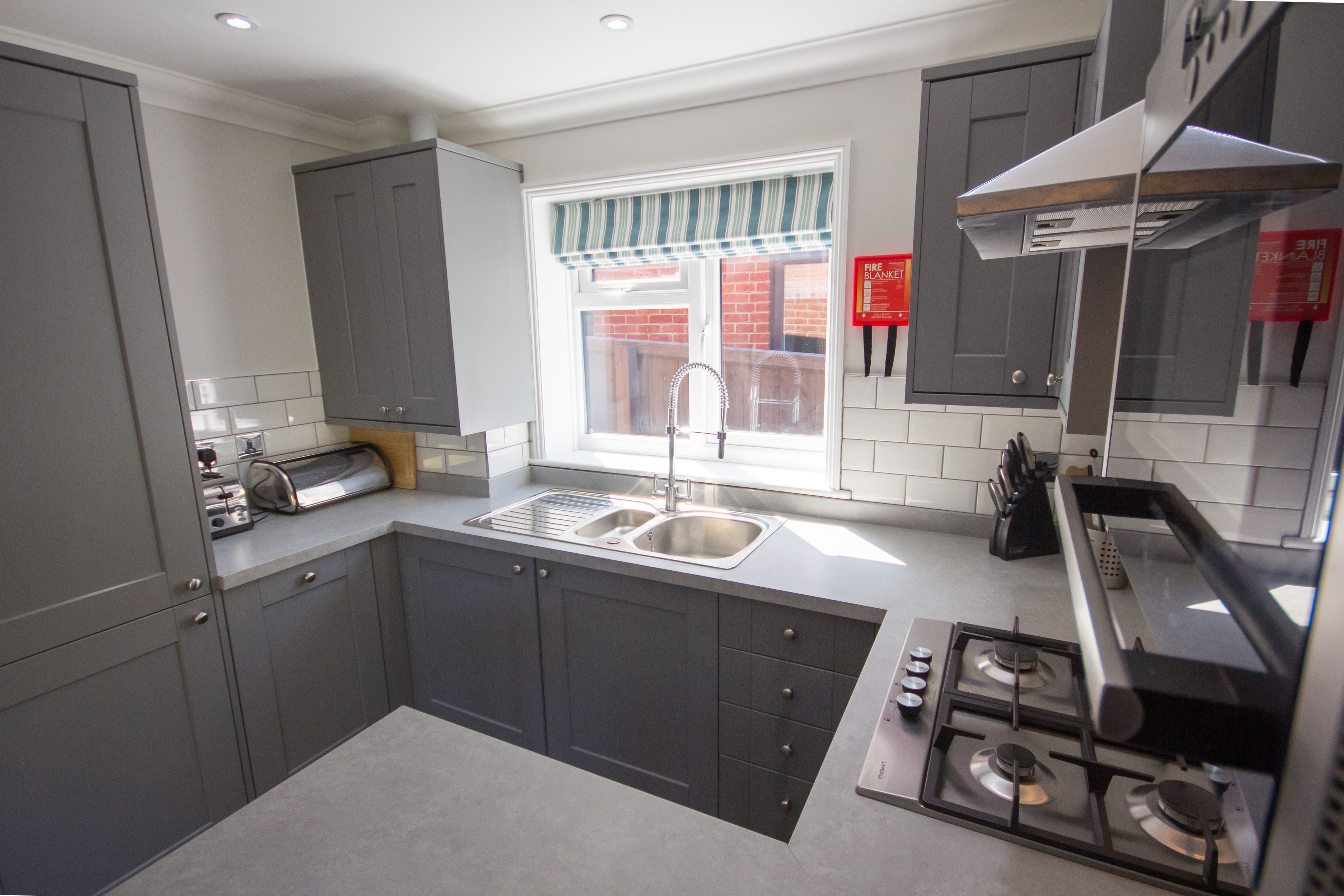 A modern kitchen with grey cabinets, a stainless steel sink, a gas stove, and a window with a striped valance.