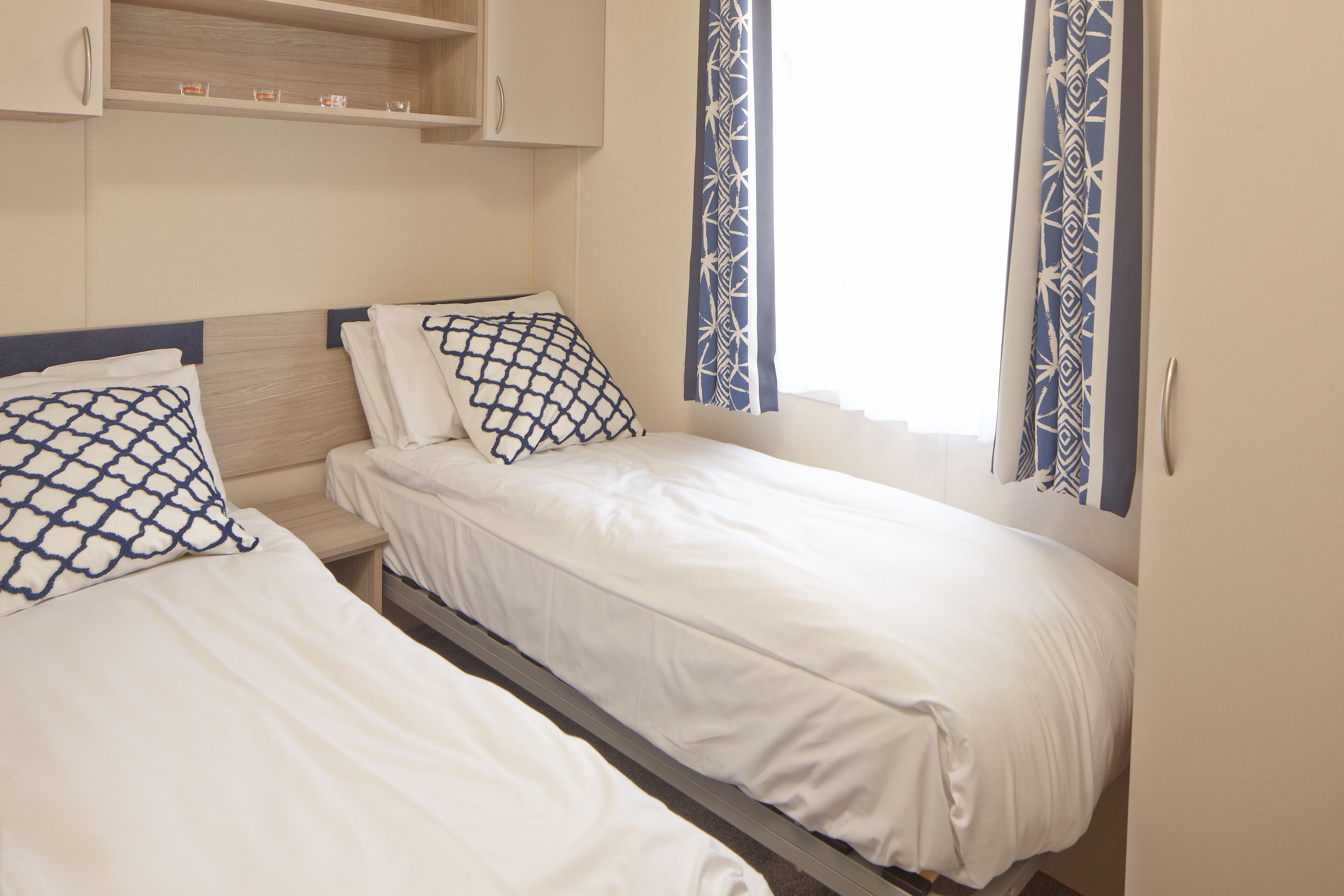 Habitación doble con dos camas, cabecero de madera, una estantería con objetos pequeños y cortinas estampadas.