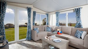 A modern living room with a beige sofa, patterned cushions, and a coffee table. There are curtains with a geometric pattern, a view of a green landscape through large windows, and a wooden floor. - St Helens Coastal Resort (Ryde)