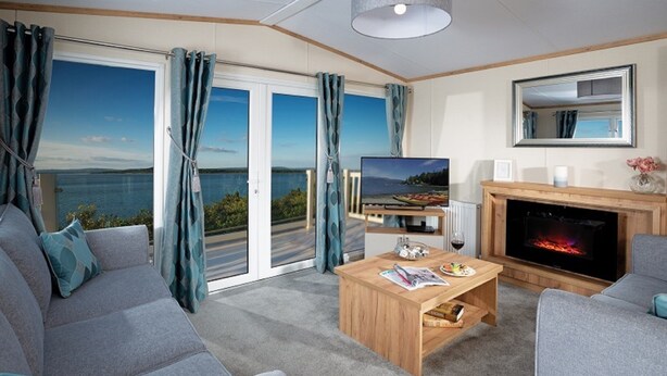 A modern living room with a sofa, coffee table, TV, fireplace, and a scenic view through the glass doors. - St Helens Coastal Resort (Ryde)