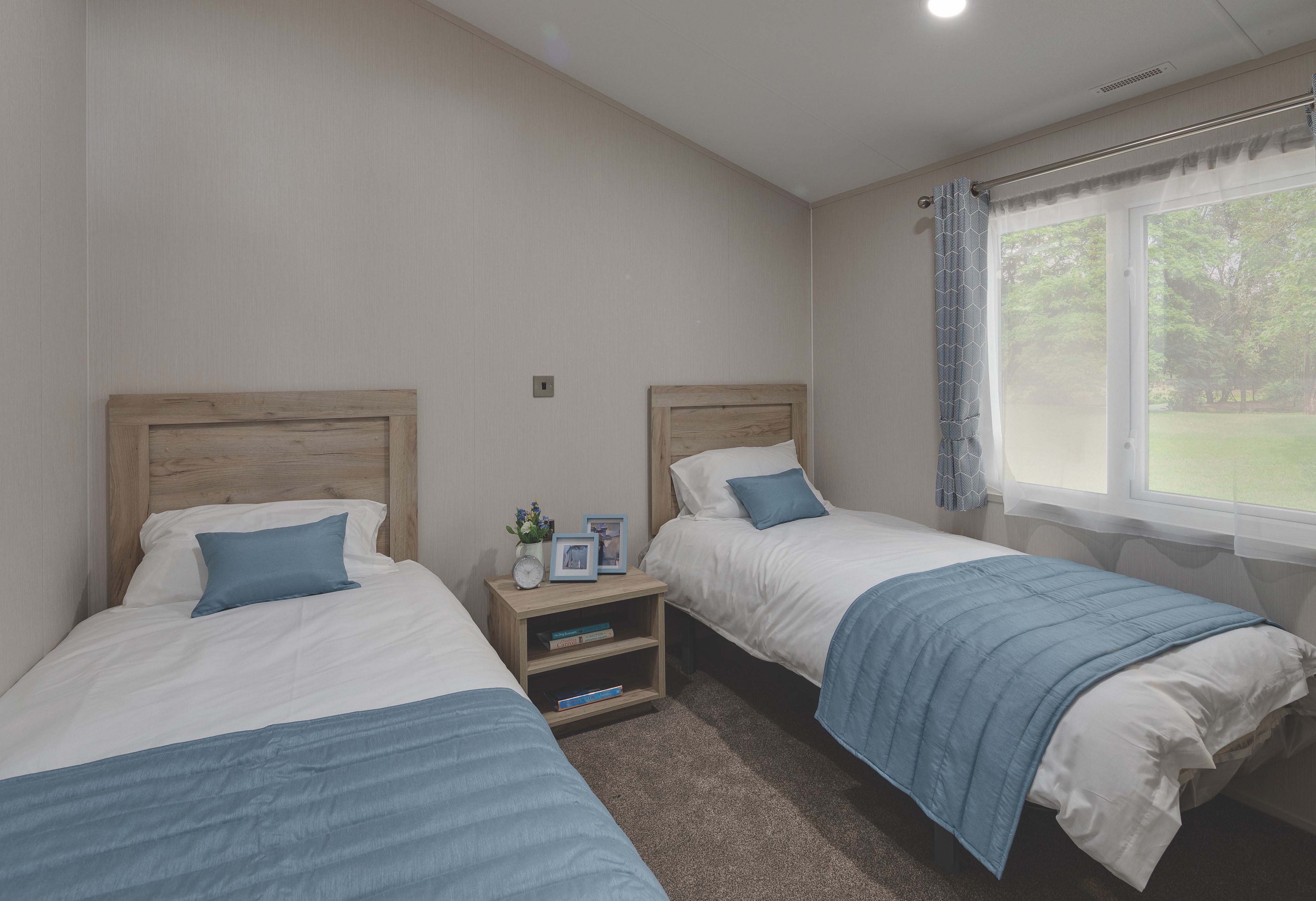 Een slaapkamer met twee eenpersoonsbedden, een nachtkastje met een klok en fotolijstjes, en een raam met uitzicht op groen.