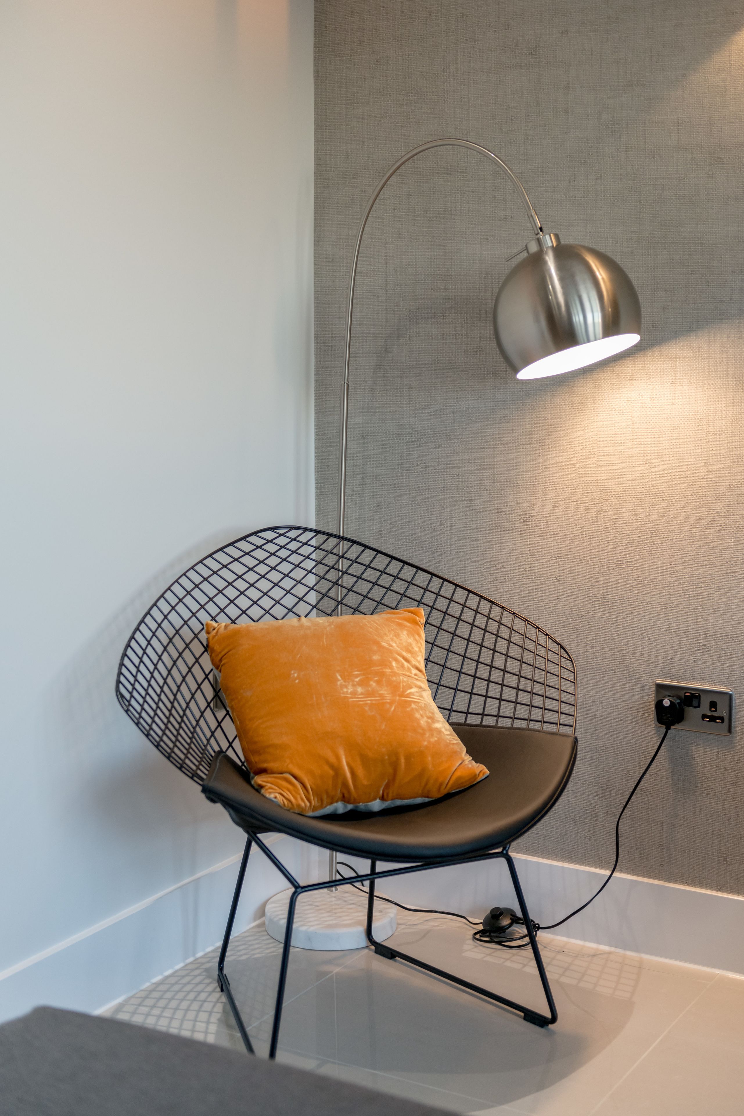 A modern chair with a wireframe backrest and a cushion, positioned against a textured wall with a power outlet nearby.