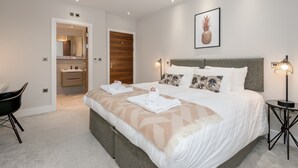 A modern bedroom with a large bed, a bedside table with a lamp, a chair, and a bathroom visible through an open door. - Silver Lining - Penthouse with parking (Edinburgh)