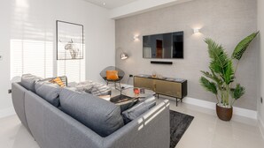 A modern living room with a grey sofa, a coffee table, a side table, a potted plant, a wall-mounted TV, and a radiator. - Silver Lining - Penthouse with parking (Edinburgh)