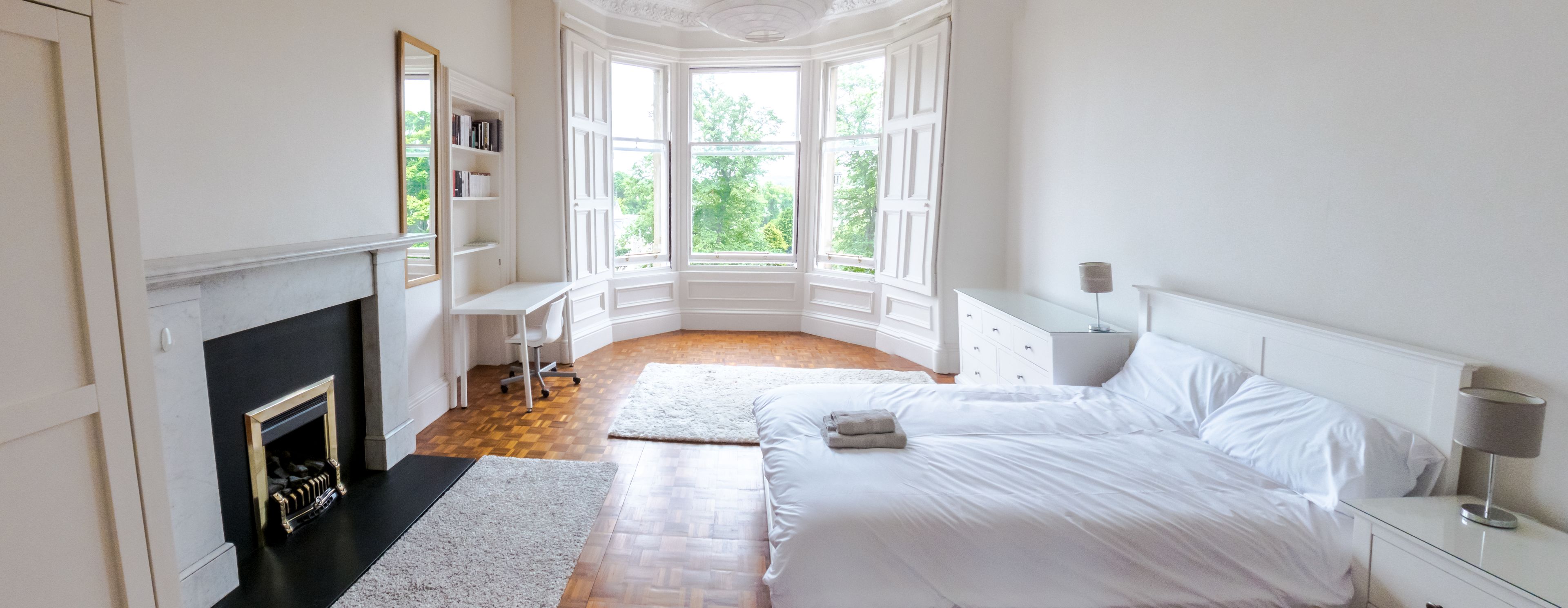 Ein Schlafzimmer mit einem großen Bett, einem Schreibtisch, einem Kamin und Blick auf Grün durch große Fenster.