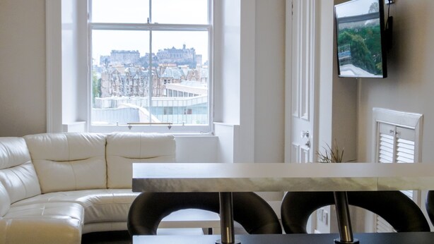 A modern living room with a white sofa, a dining table with chairs, and a large window offering a city view. - Silver Lining - Meadows Apartments (Edinburgh)