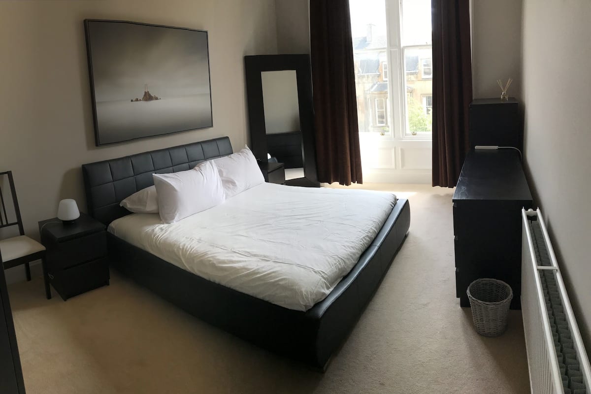 Une chambre d’hôtel moderne avec un grand lit, une table de chevet avec une lampe, une commode, un miroir et un tableau au mur.