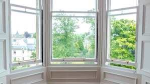 A corner room with three large windows, a wooden floor, and a view of buildings and trees. - Silver Lining - Meadows Apartments (Edinburgh)