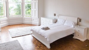 A bedroom with a large window, a bed, a nightstand, and a dresser. - Silver Lining - Meadows Apartments (Edinburgh)