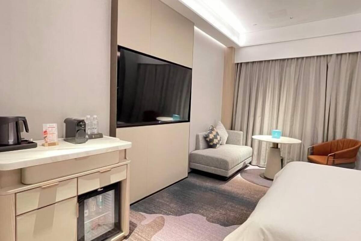 Ein Hotelzimmer mit Bett, kleinem Schreibtisch mit Kaffeemaschine, Stuhl, Sofa und Fernseher.
