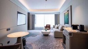 A modern hotel room with a sofa, a round coffee table, a desk with a computer, and a large window with curtains.