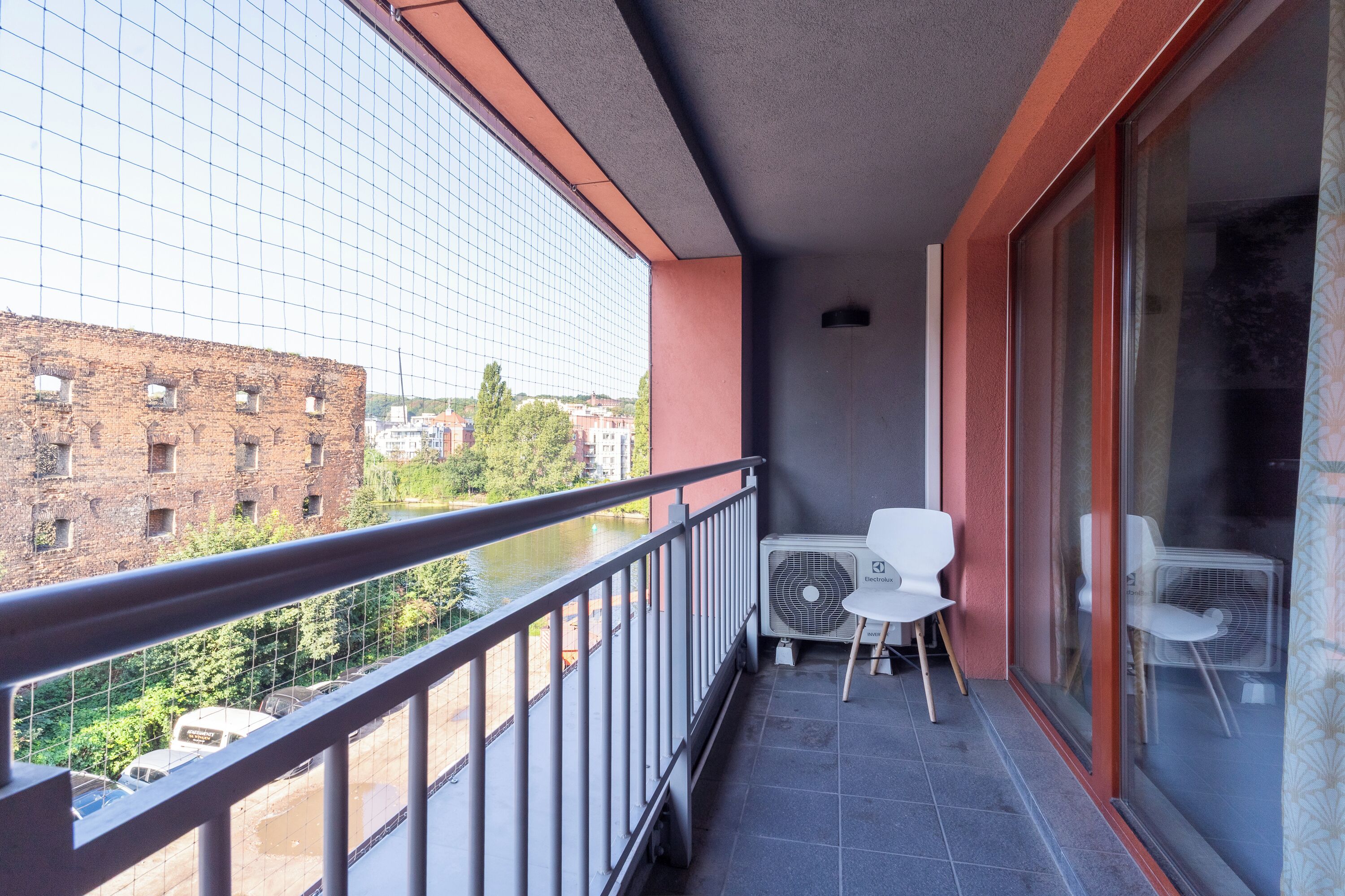 A balcony with a view of a river, brick buildings, and a modern glass building. There are two white chairs and an air conditioning unit.