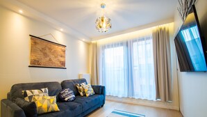 A modern living room with a dark blue sofa, patterned cushions, a wooden floor, and a large window with sheer curtains.