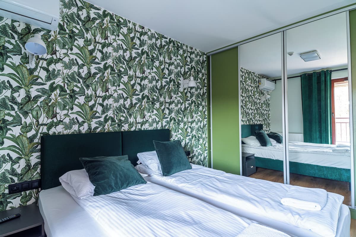 Una cama bien tendida con sábanas blancas y almohadas verdes, una mesita de noche con un control remoto, y una pared con estampado de hojas.