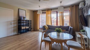 A modern living room with a sofa, a dining table, and chairs. There is a bookshelf, a potted plant, and a TV mounted on the wall.
