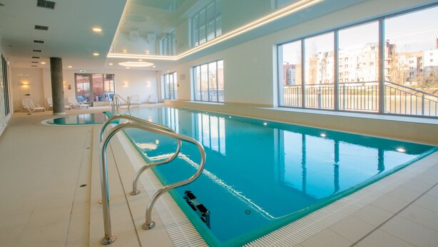 An indoor swimming pool with clear glass walls, a metal pool ladder, and a view of buildings outside.