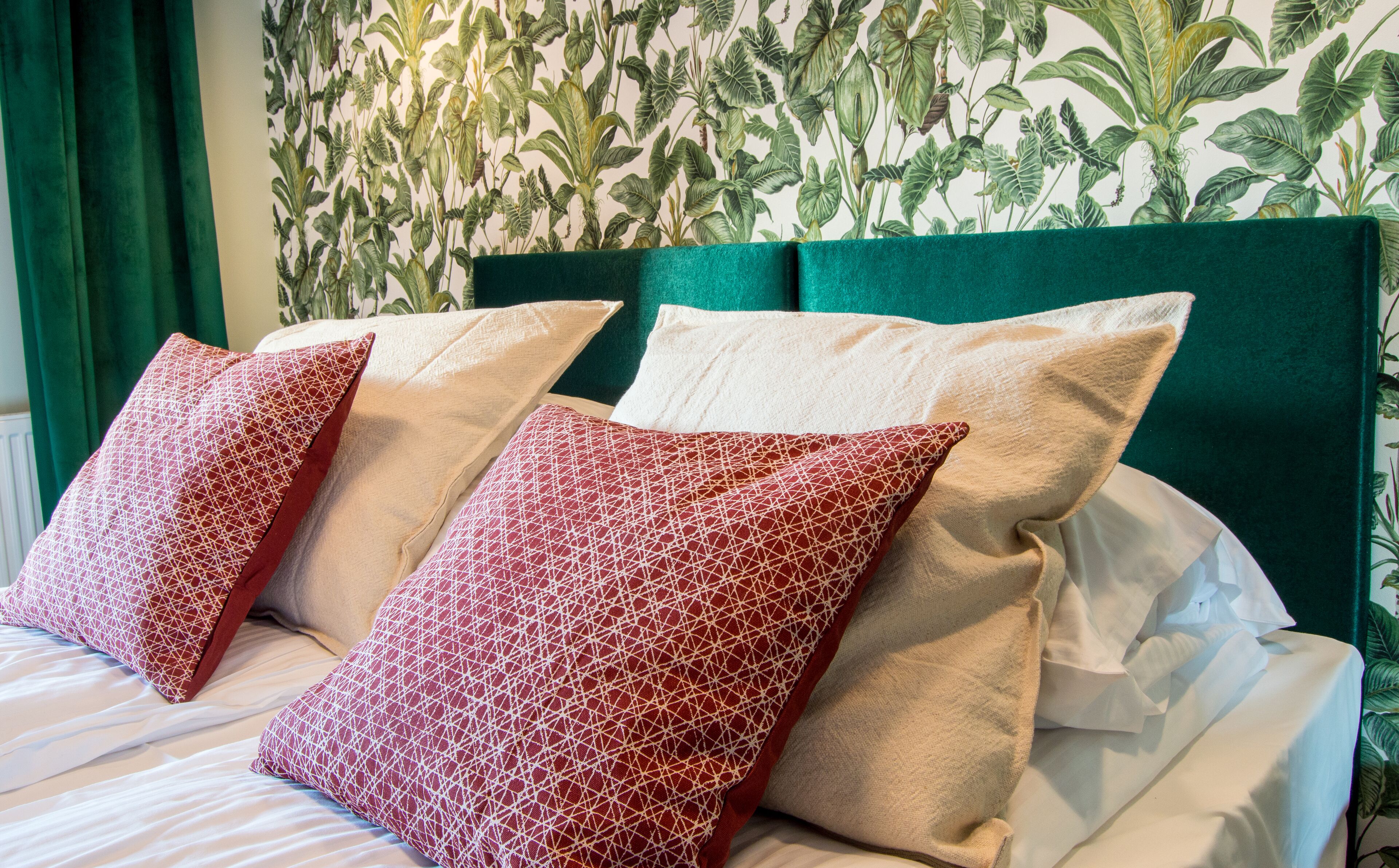 Un lit avec des oreillers à motifs rouges et blancs, une tête de lit verte et un mur recouvert d’un papier peint à motifs de feuilles.