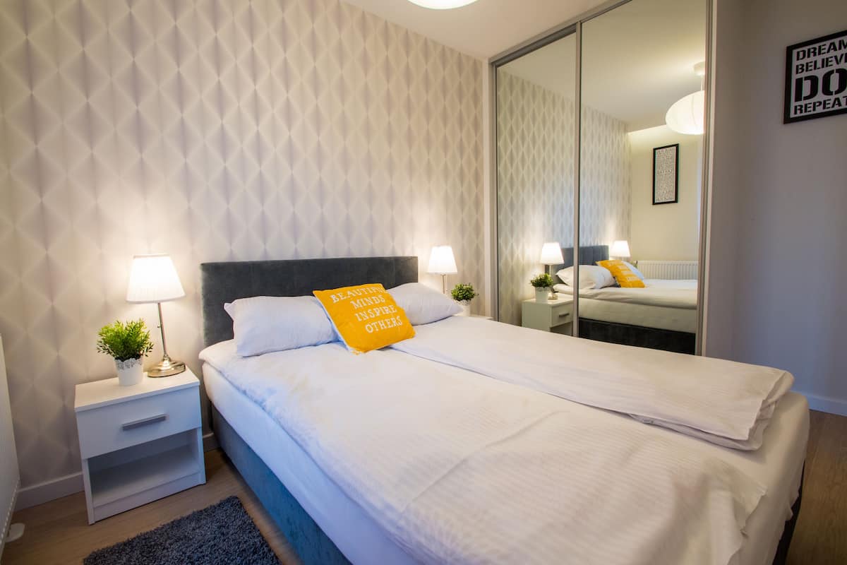 Una cama bien hecha con sábanas blancas y una almohada amarilla con texto, una mesita de noche con una lámpara y un espejo grande que refleja la habitación.