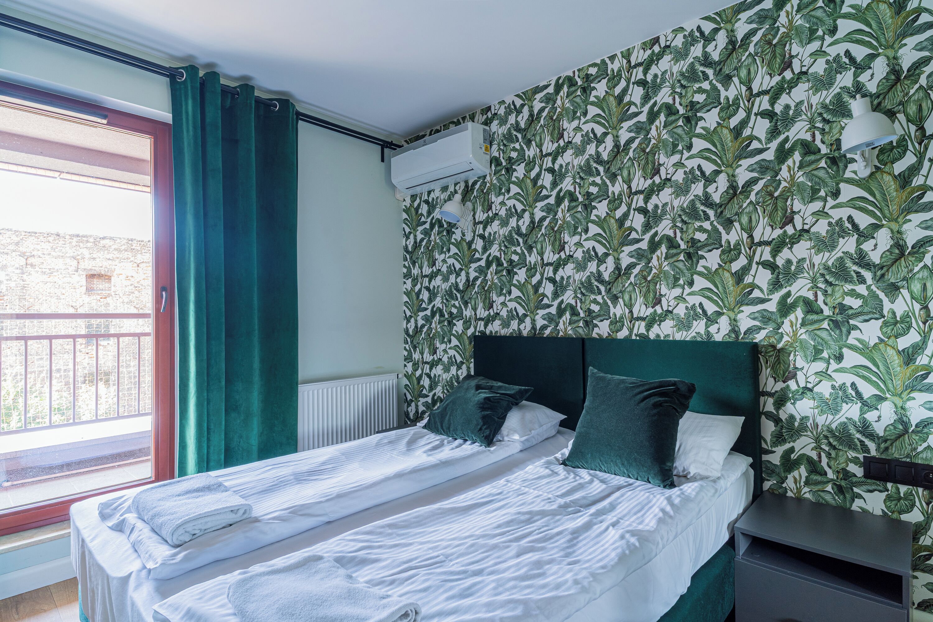 A double bed with white linens and green pillows, a window with a view of a building, and a wall with a leafy pattern.