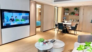 Moderni hotellihuone, jossa on taulutelevisio, ruokailutila pöytineen ja tuoleineen sekä hyllyllinen kasveja.