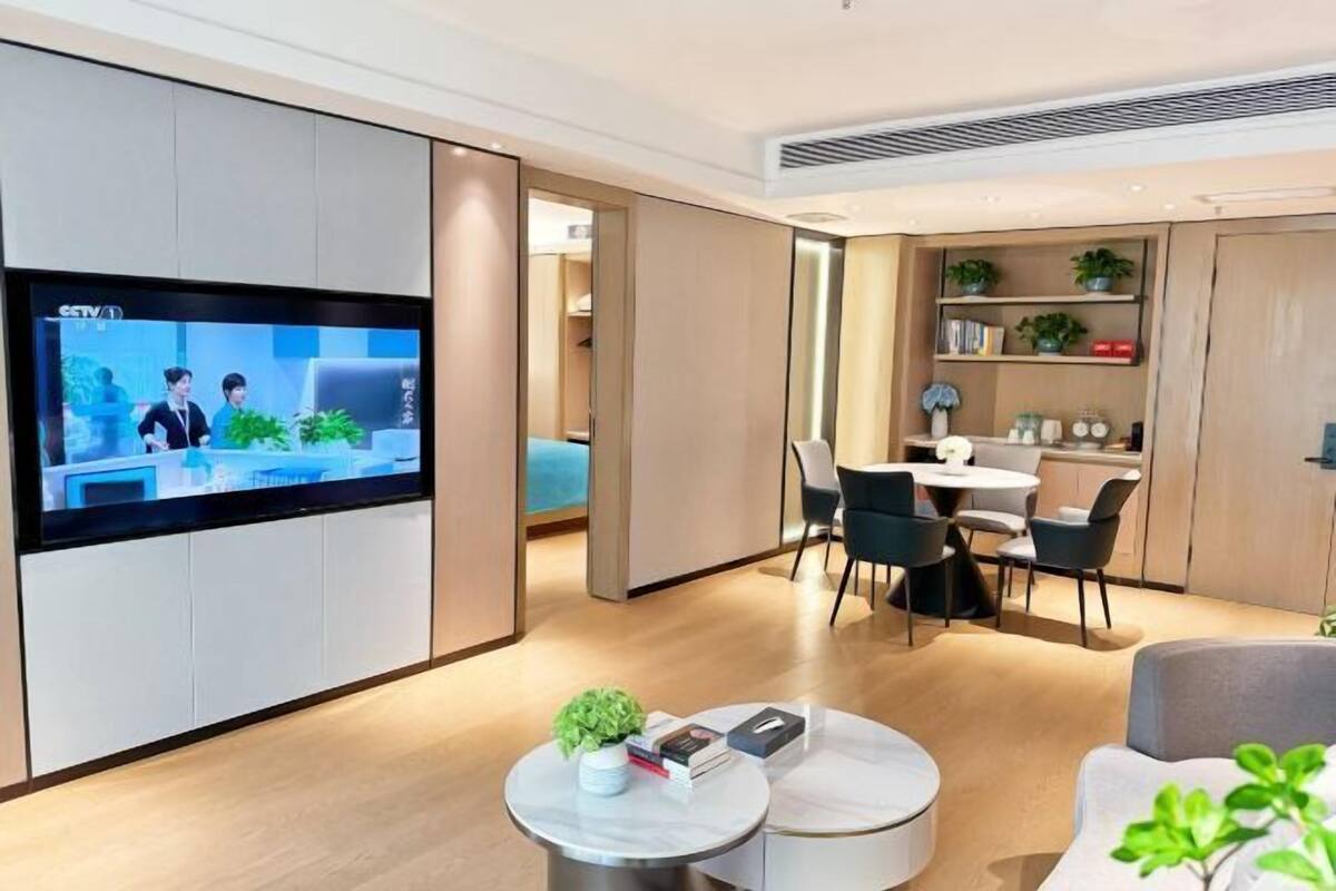 Une chambre d’hôtel moderne dotée d’une télévision à écran plat, d’un coin repas avec une table et des chaises, et d’une bibliothèque agrémentée de plantes.