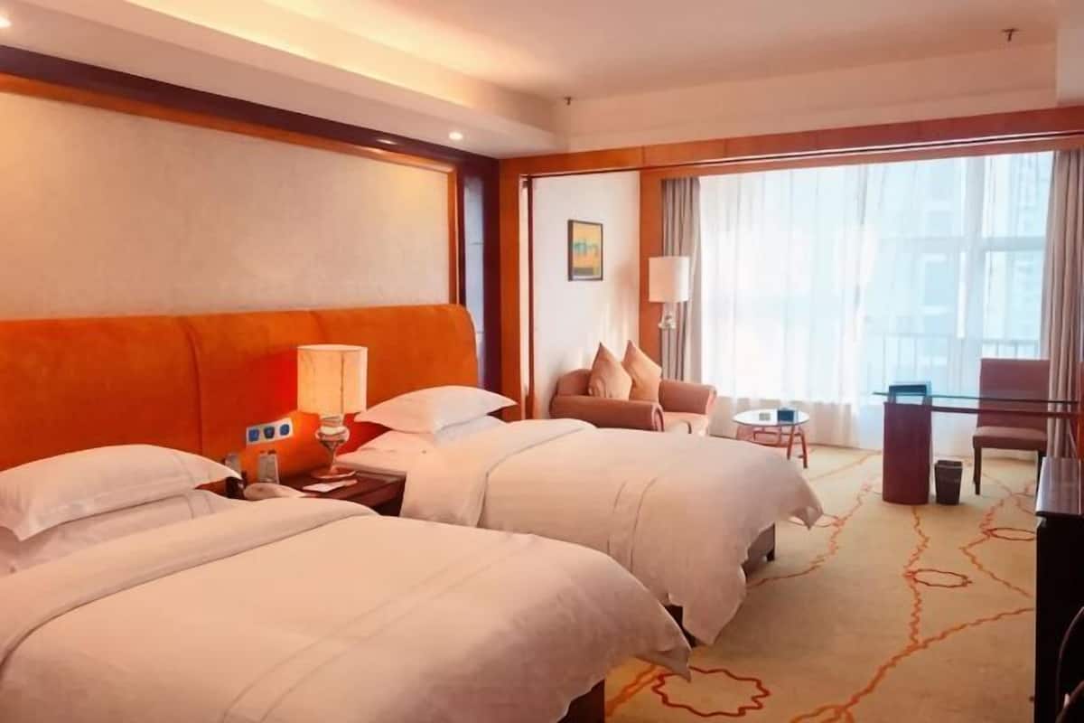 A hotel room with two beds, a desk, a chair, a lamp, and a view of the outdoors.